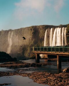 A breathtaking view of waterfalls cascading beside a viewing deck with a bird flying in the blue sky.