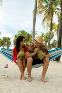 A couple enjoys coconut drinks in a hammock under palm trees on a tropical beach.